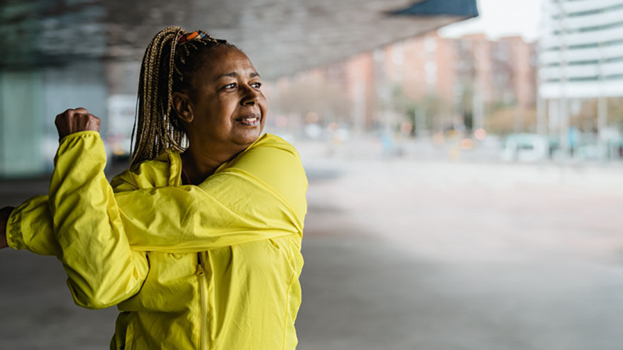 Mulher na menopausa a realizar alongamentos nos braços, em ambiente urbano e vestida com casaco amarelo