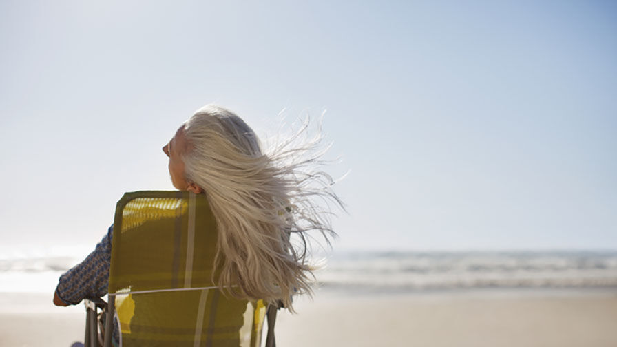 Mulher com cabelo longo e grisalho sentada numa cadeira à beira-mar 