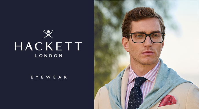 Homem com óculos Hackett London, camisa às riscas, gravata azul e casaco bege com lenço vermelho.
