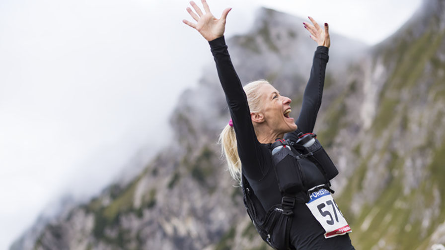 Mulher na menopausa durante uma corrida em ambiente montanhoso, a celebrar com os braços levantados, representando a superação do cansaço 